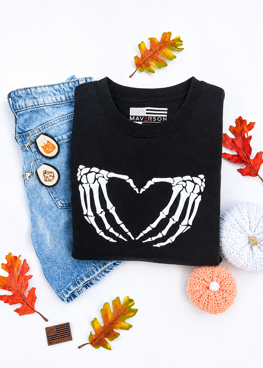 Black sweatshirt with skeleton hands forming a heart design, paired with denim jeans and autumn leaves on a white background.