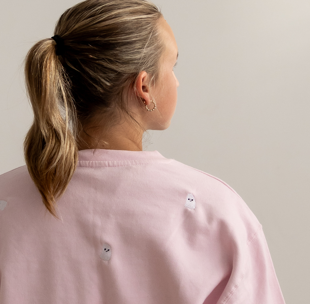 A woman shown from the back wearing a light pink crewneck sweatshirt with small embroidered ghost details, set against a plain light gray background.