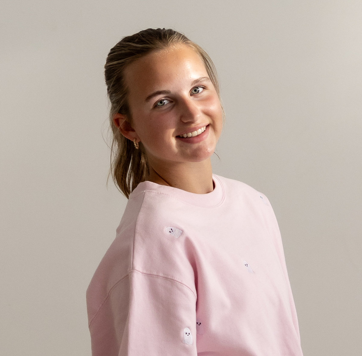 A smiling young woman with blonde hair tied back, wearing a light pink crewneck sweatshirt with small embroidered cartoon face details, photographed against a plain light gray background.