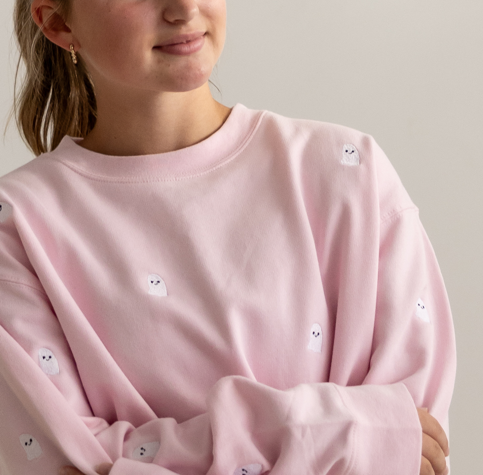 A close-up of a person wearing a light pink crewneck sweatshirt with small white embroidered ghost motifs scattered across the front, set against a plain light gray background.
