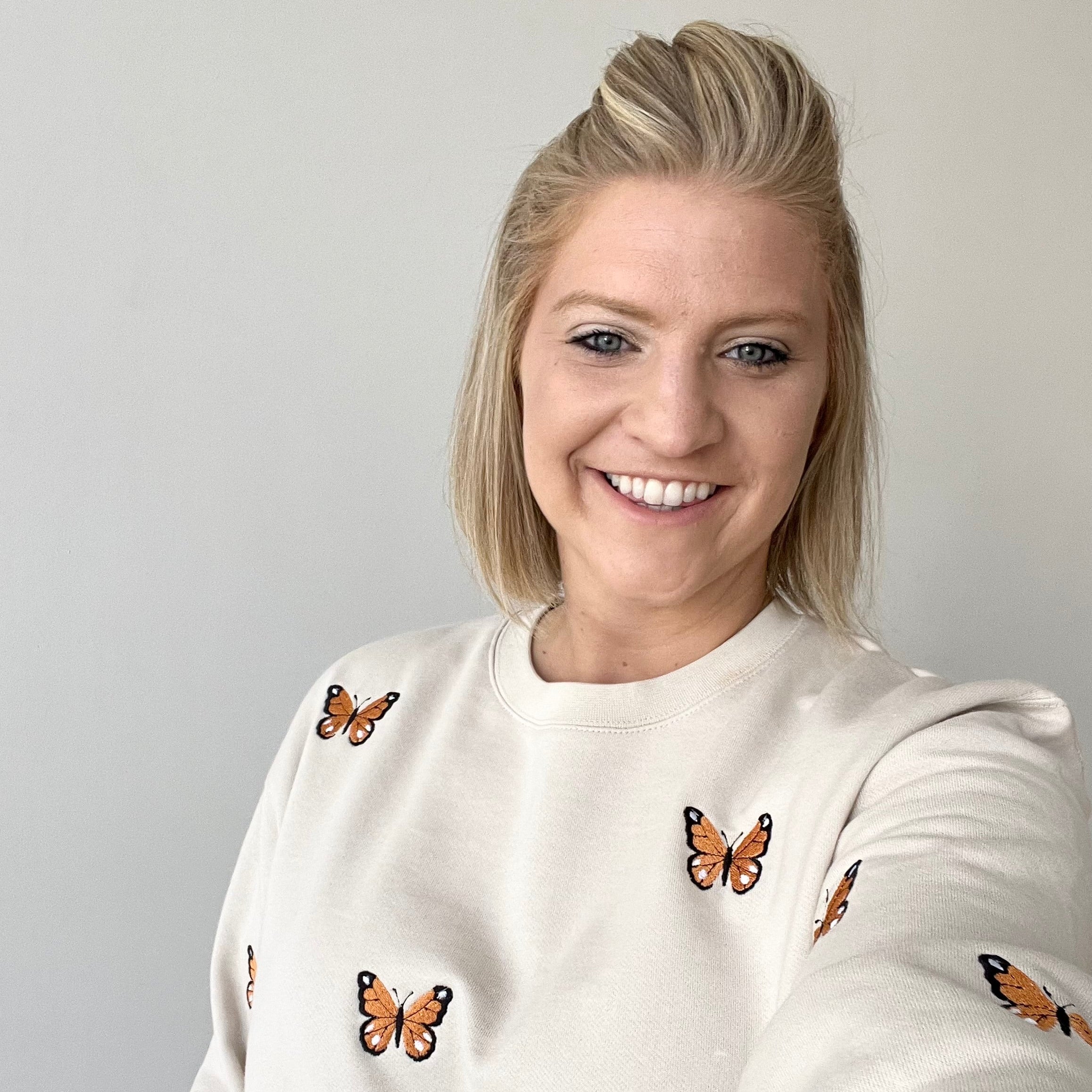 A smiling blonde woman wearing a cream-colored crewneck sweatshirt with embroidered monarch butterflies, taking a selfie against a plain light gray wall.