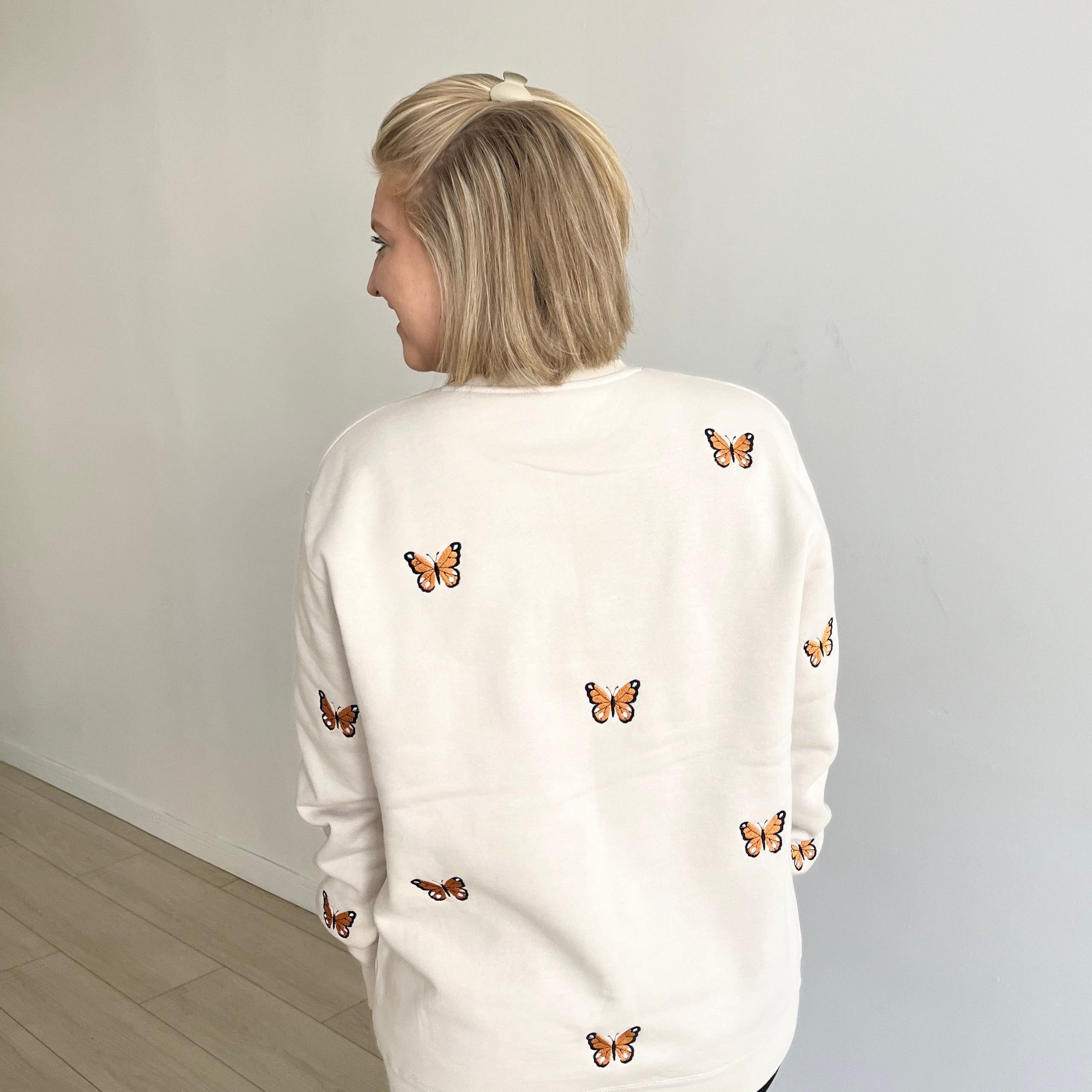 A woman with blonde hair is shown from behind wearing a cream-colored crewneck sweatshirt decorated with small embroidered butterflies, paired with black pants, standing in a minimalist room with a white wall and light wood floor.