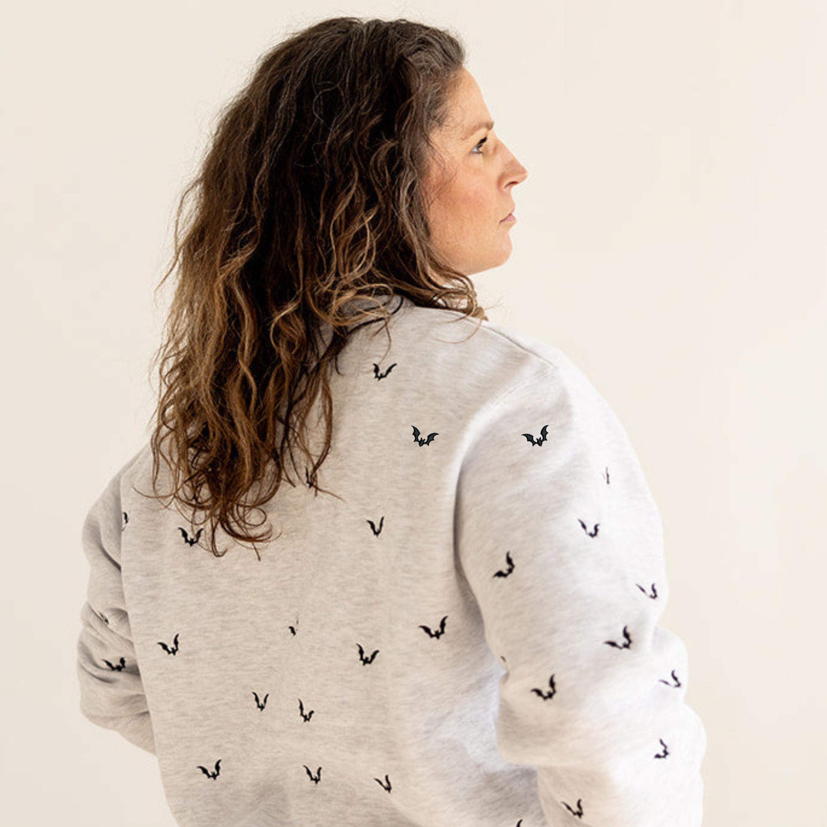 A woman with long, wavy brown hair is shown from the back wearing a light heather gray sweatshirt with a repeating black embroidered logo, paired with dark blue jeans, against a plain light beige background.