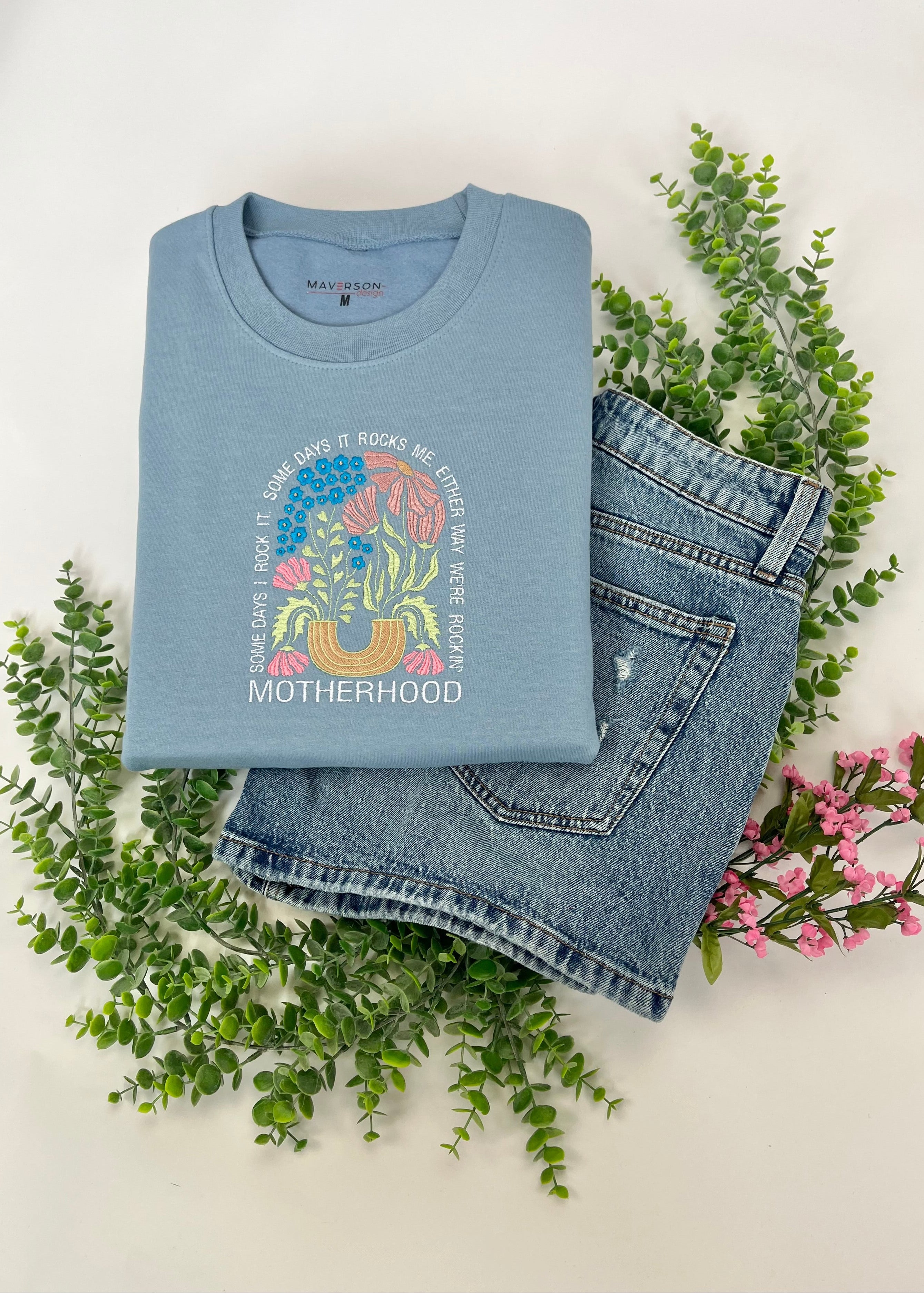 Folded baby blue crewneck sweatshirt with 'Motherhood' embroidery and jeans shorts on a white background with greenery and flowers.