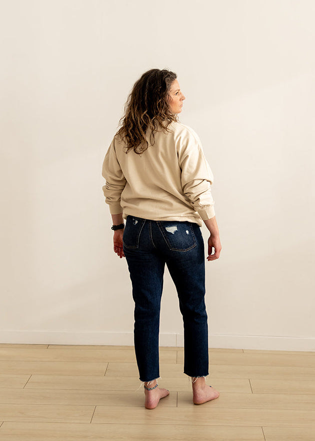 A woman stands with her back to the camera in a minimalist room with a beige wall and light wood floor, wearing a beige sweatshirt, dark blue jeans, and pink shoes.