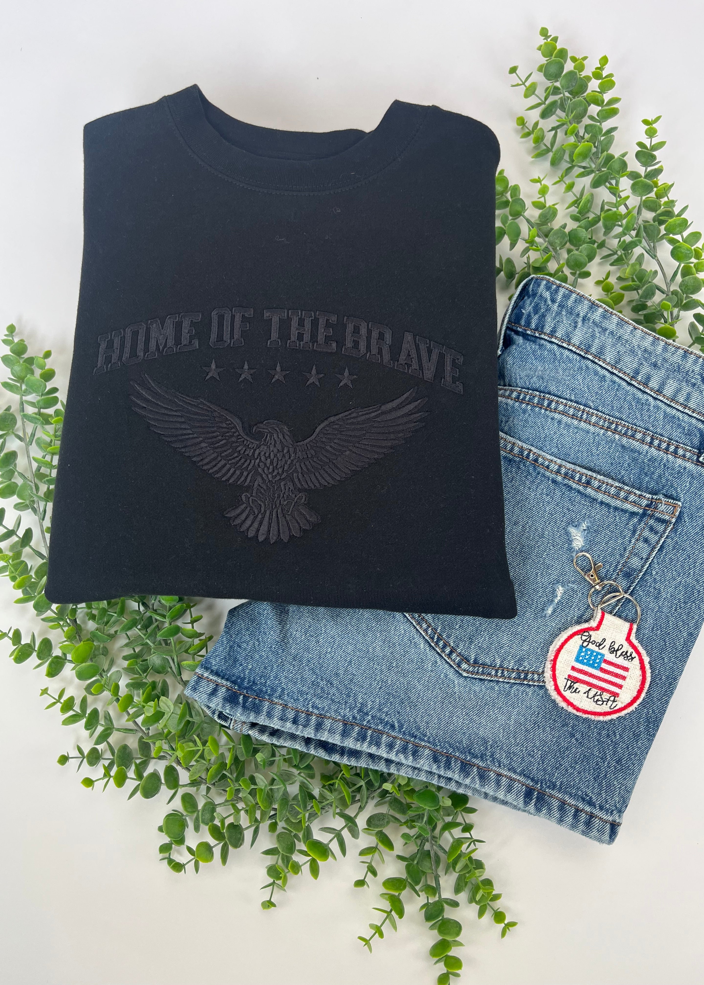 Black sweatshirt with eagle design and text, paired with blue jeans and a keychain on a white background with greenery.