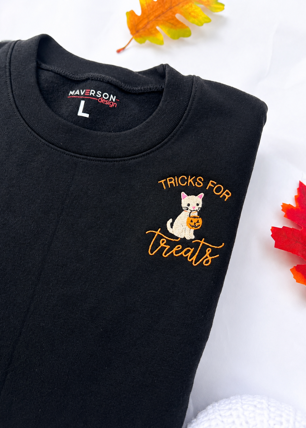 Black sweatshirt with Halloween design and text, brand 'Maverson' visible, surrounded by autumn leaves- close up