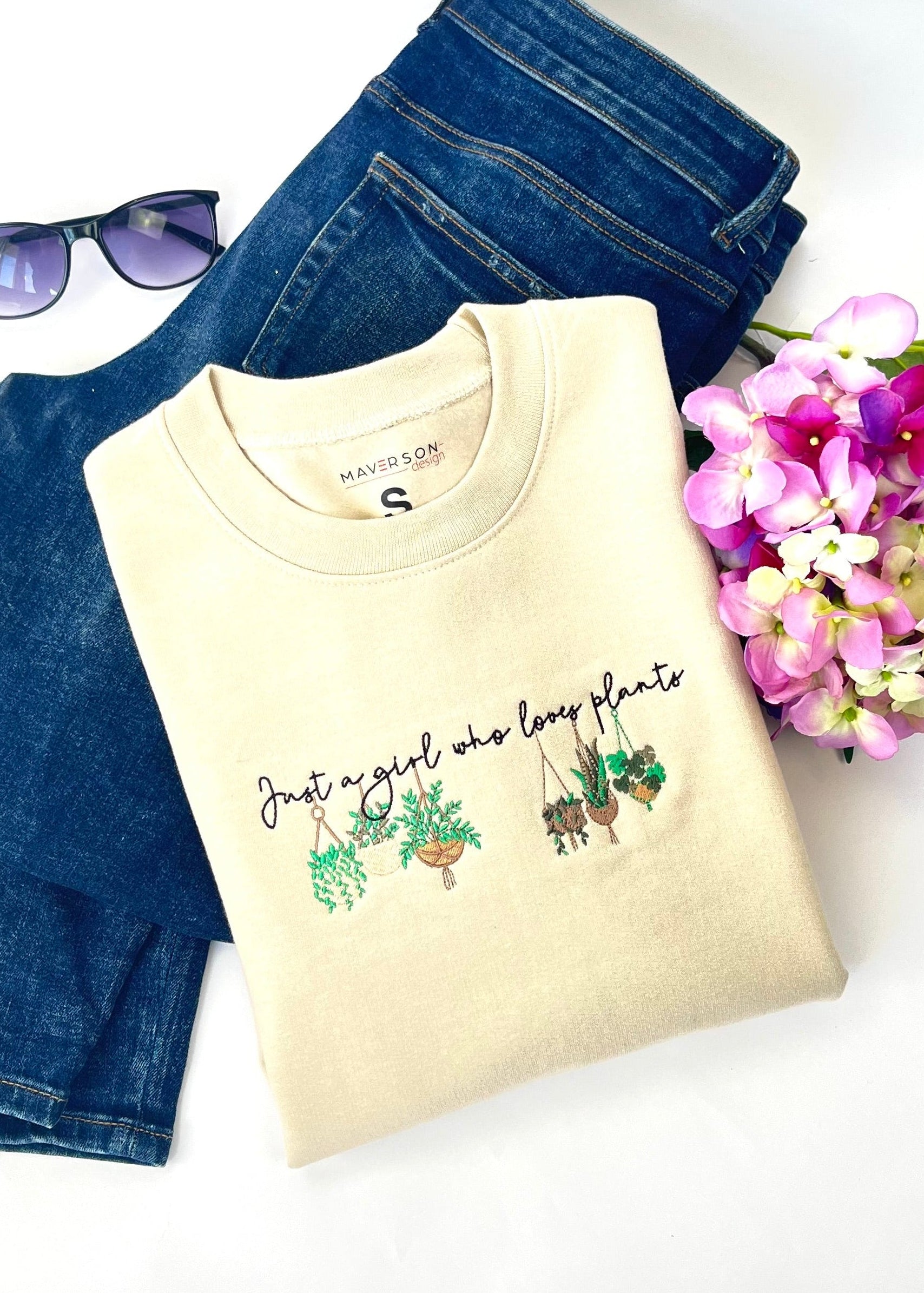 Beige crewneck sweatshirt with plant-themed embroidery design and text, placed on a white surface with jeans and flowers.