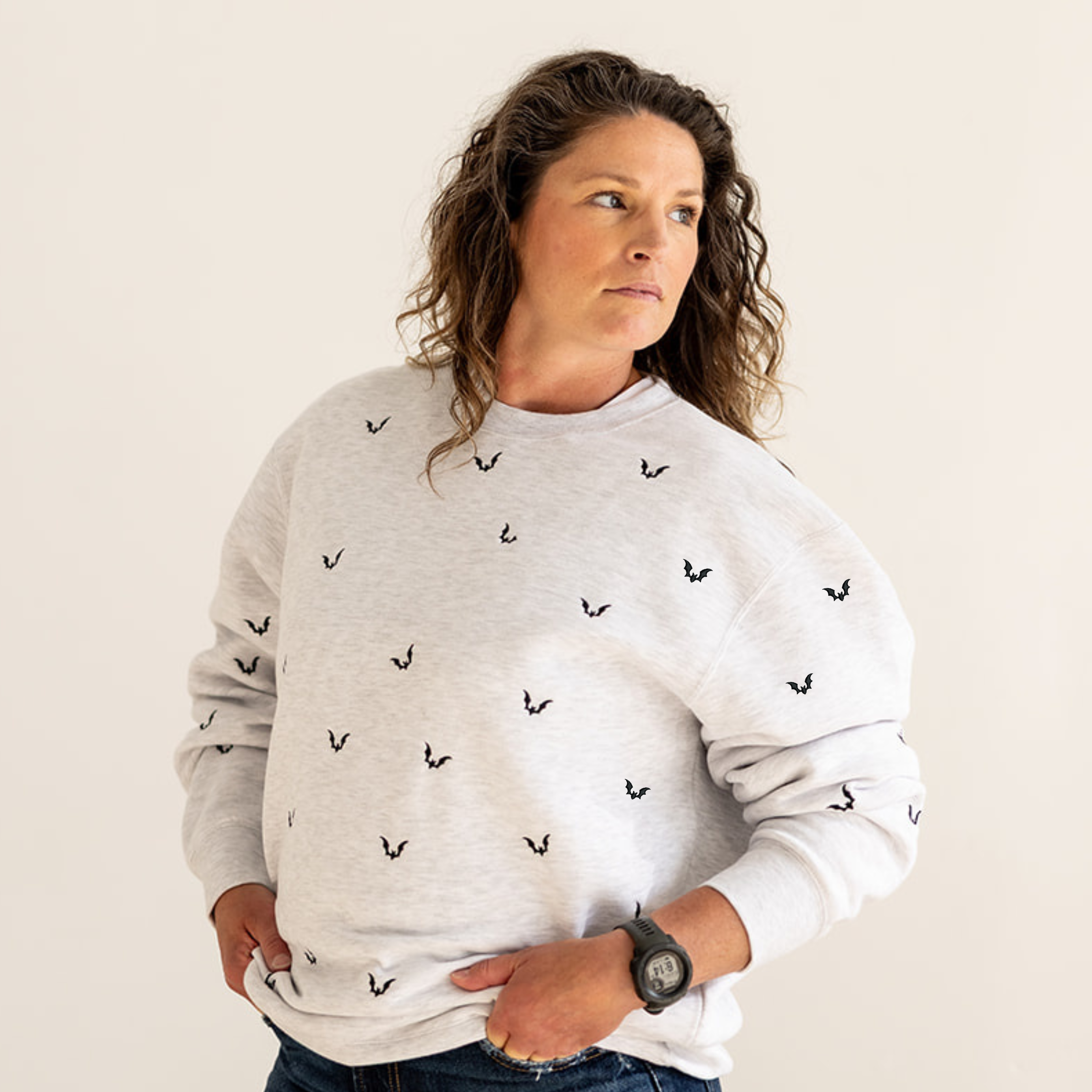 A woman wearing a light gray crewneck sweatshirt with a small black embroidered pattern, paired with dark blue jeans, standing against a plain beige background.