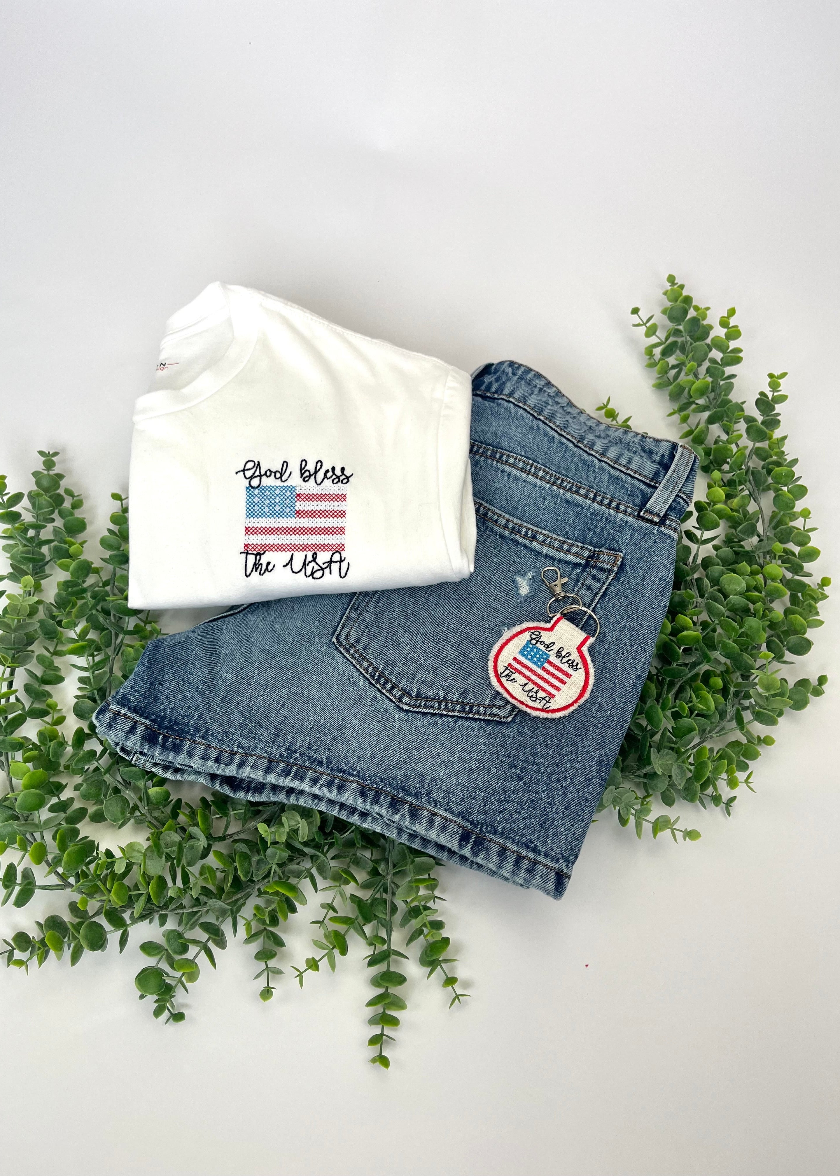 White t-shirt with 'God bless the USA' text and blue jeans with American flag patch on a white background with greenery.