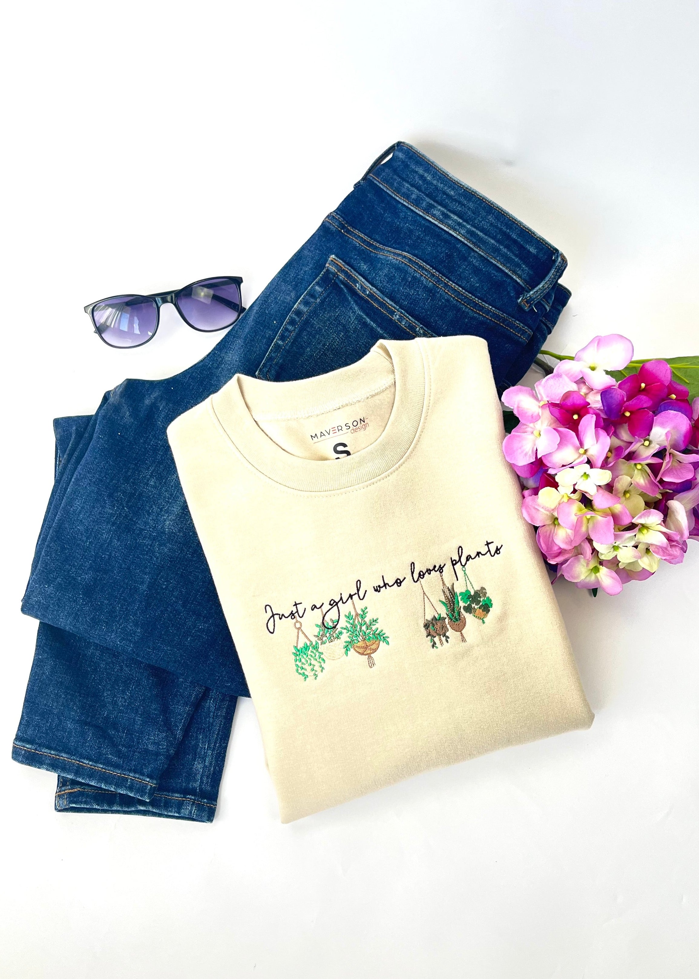 Beige crewneck with plant-themed embroidery design, blue jeans, sunglasses, and flowers on a white background