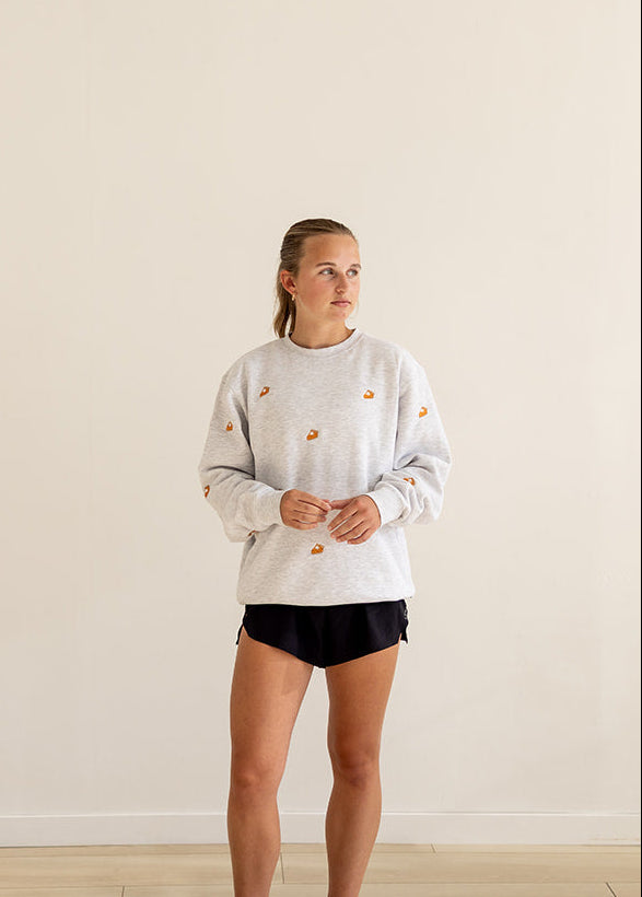 A woman stands in a minimalist room with a cream wall and light wood floor, wearing a white crewneck sweatshirt with small slice of pie embroidered details, black athletic shorts, and white sneakers.