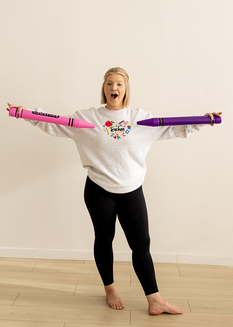 A woman stands barefoot on a light wood floor against a plain off-white wall, holding a pink and purple toy crayon-like exercise bar across her outstretched arms. She is wearing a white sweatshirt with a colorful graphic and black leggings.