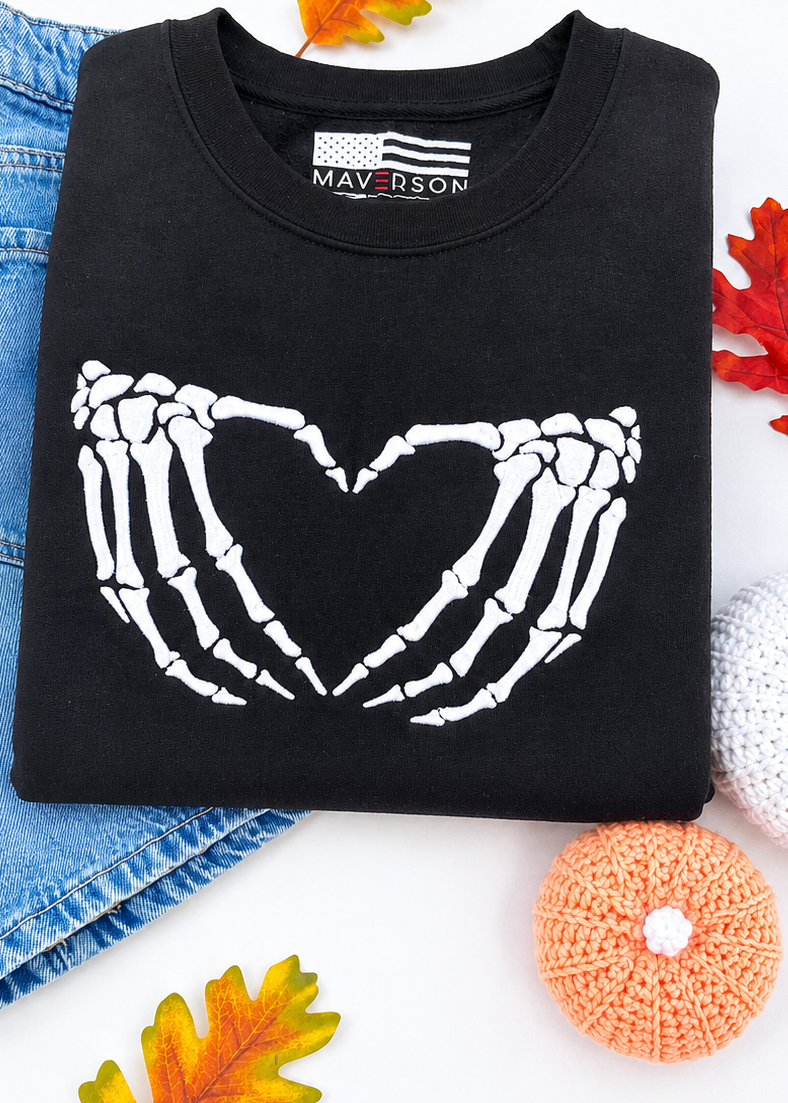 Black sweatshirt with skeleton hands forming a heart design, placed on denim shorts with autumn leaves and decorative items.