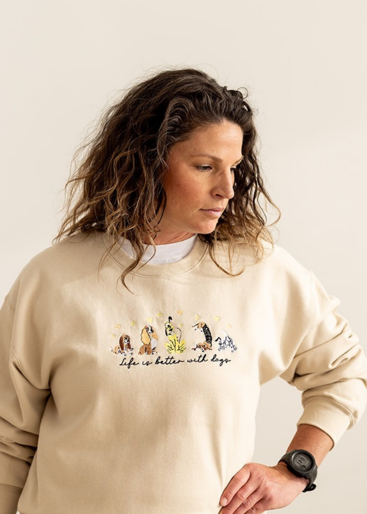 A woman wearing a cream-colored crewneck sweatshirt with a graphic of cartoon dogs and the text 'Life is better with dogs' paired with dark blue jeans, set against a plain beige background.
