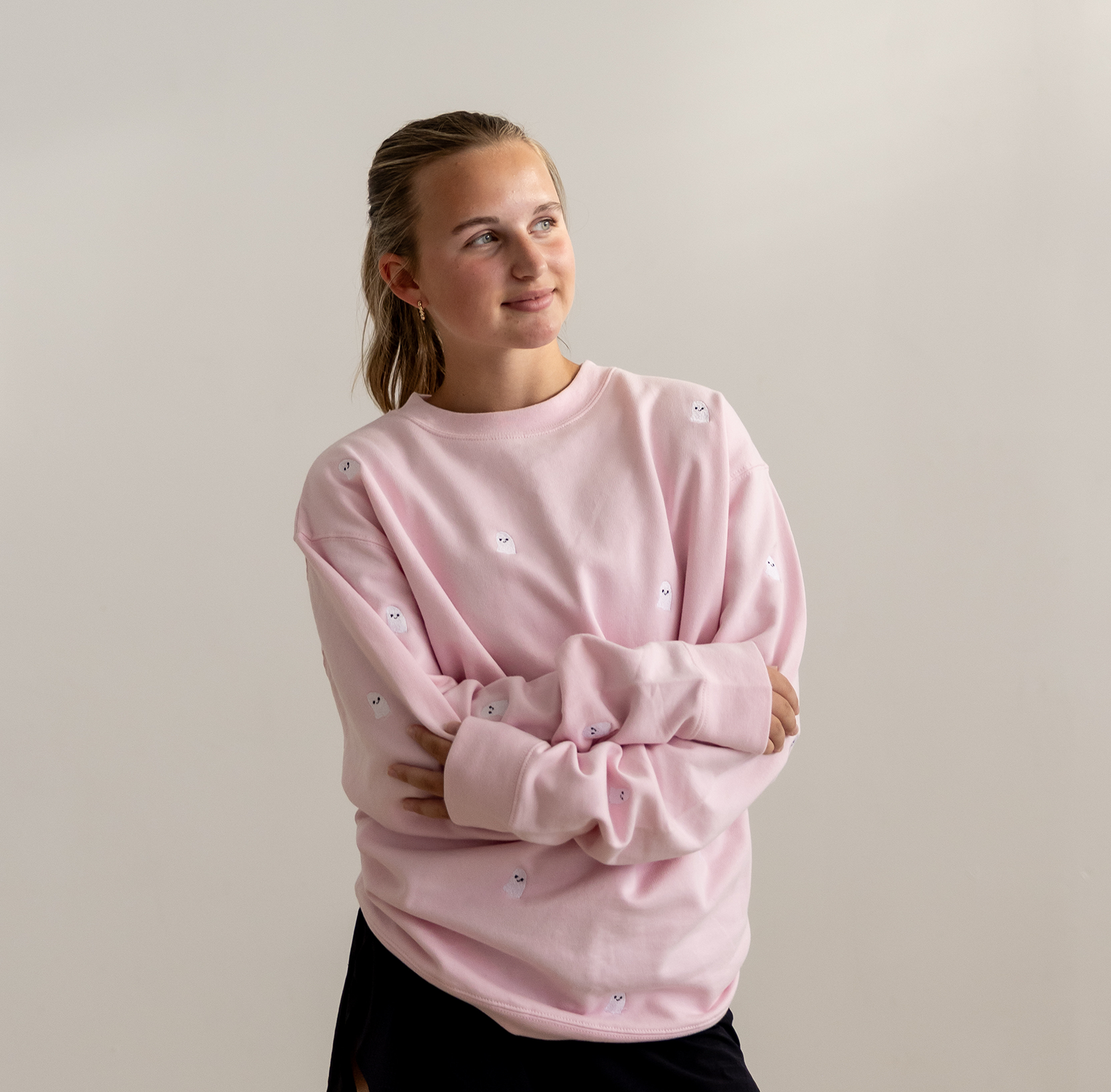A young woman with her hair in a ponytail wearing a light pink crewneck sweatshirt with small embroidered smiley faces, posed against a neutral light gray background.