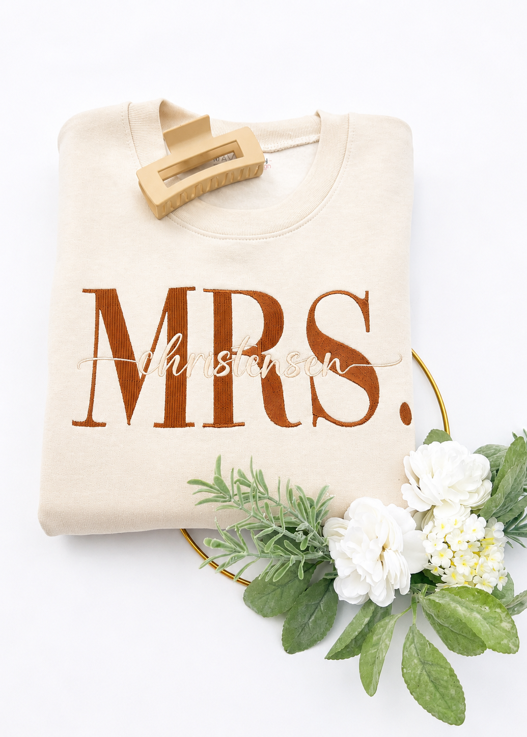 Beige sweatshirt with 'MRS.' printed on it, hanging on a white background with flowers and greenery.