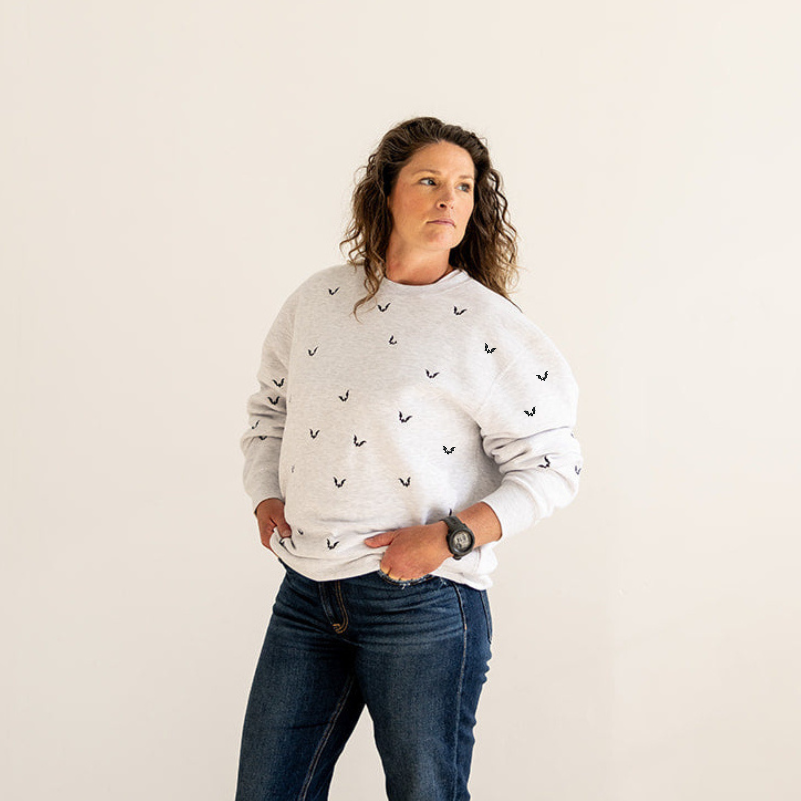 A woman stands barefoot in a minimalist room with light wood floors and a cream wall, wearing a white crewneck sweatshirt with a small black embroidered pattern and dark blue jeans.