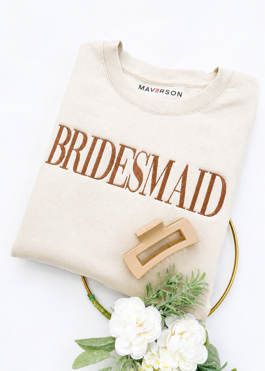 Beige sweatshirt with 'BRIDESMAID' text, wooden ring, and flowers on a white background