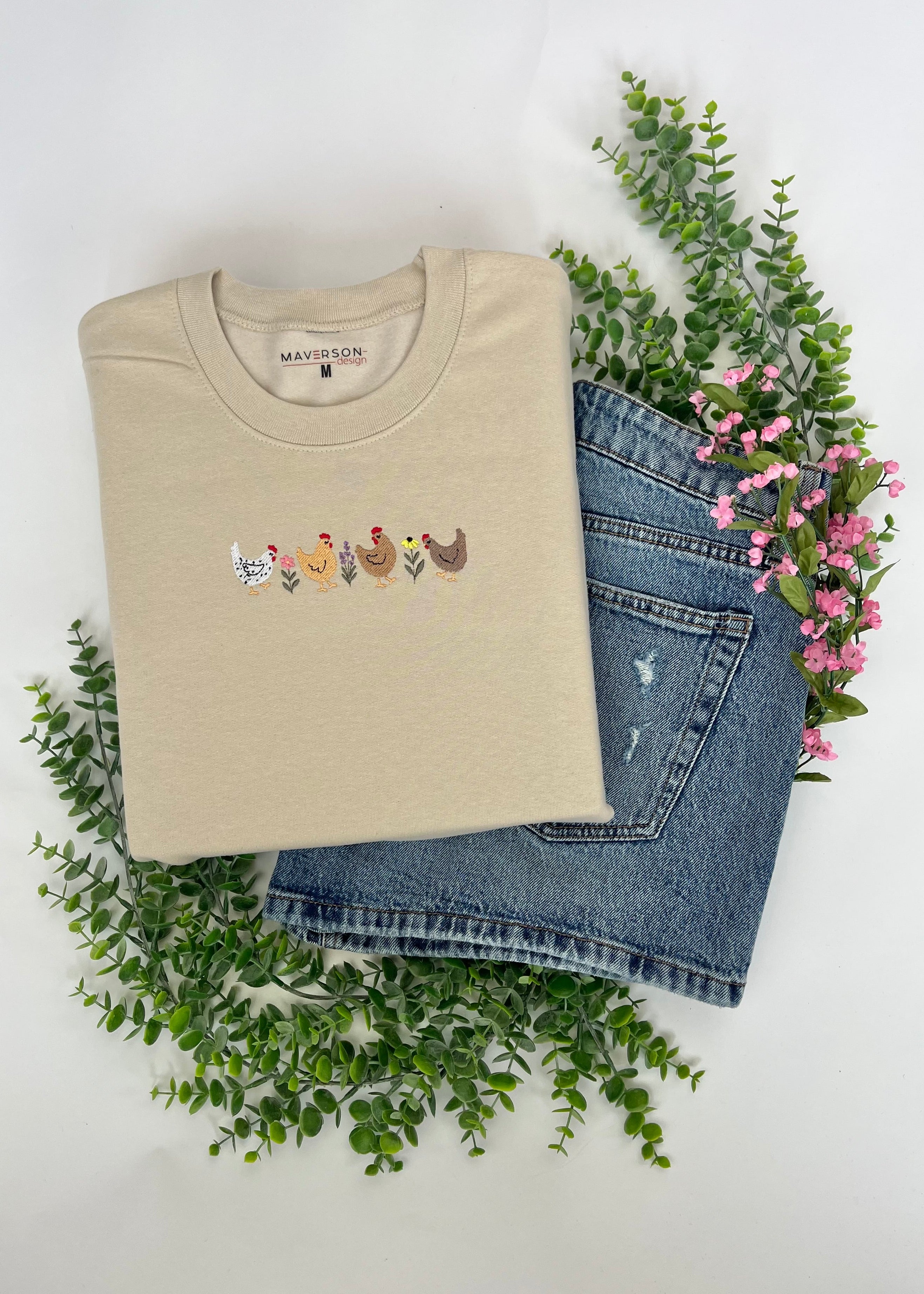Beige sweatshirt with embroidered design and blue jeans on a white background with greenery and flowers.