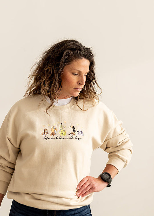 A woman wearing a cream-colored crewneck sweatshirt with embroidered  design on the chest, paired with dark blue jeans, standing against a plain off-white wall.