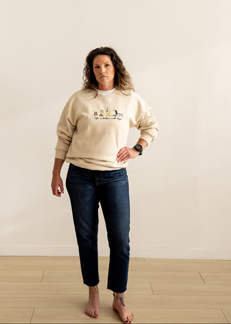 A woman stands barefoot in a minimalist studio setting with a warm beige wall and light wood floor, wearing a cream-colored crewneck sweatshirt with a colorful embroidered and dark blue jeans.