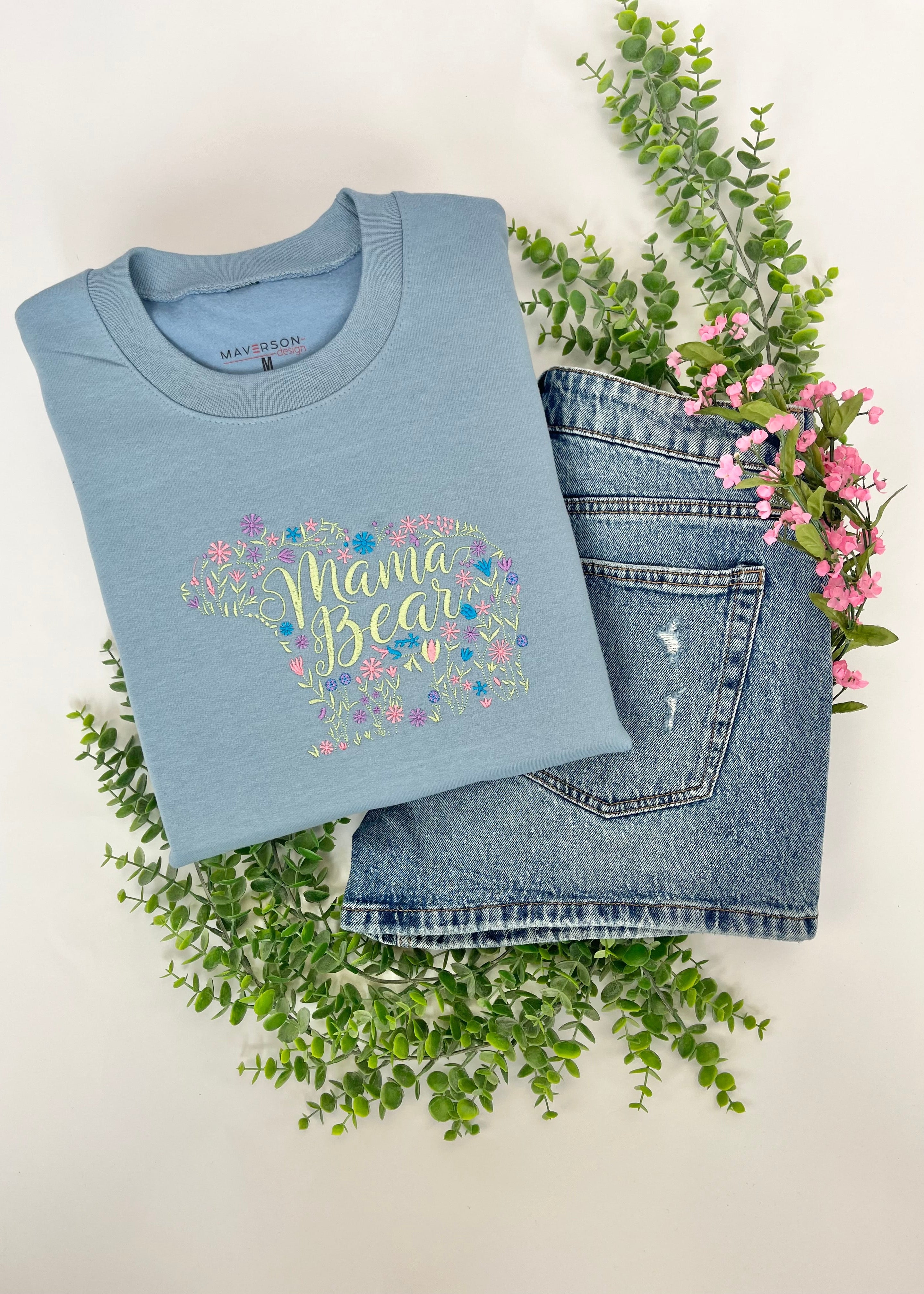 Light blue crewneck sweatshirt with 'Mama Bear' embroidery design on a white background with greenery and flowers.