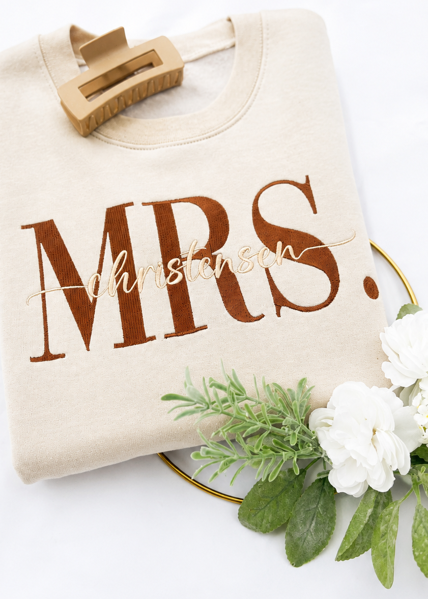 Beige tote bag with 'MRS.' text, wooden clip, and white flowers on a white background