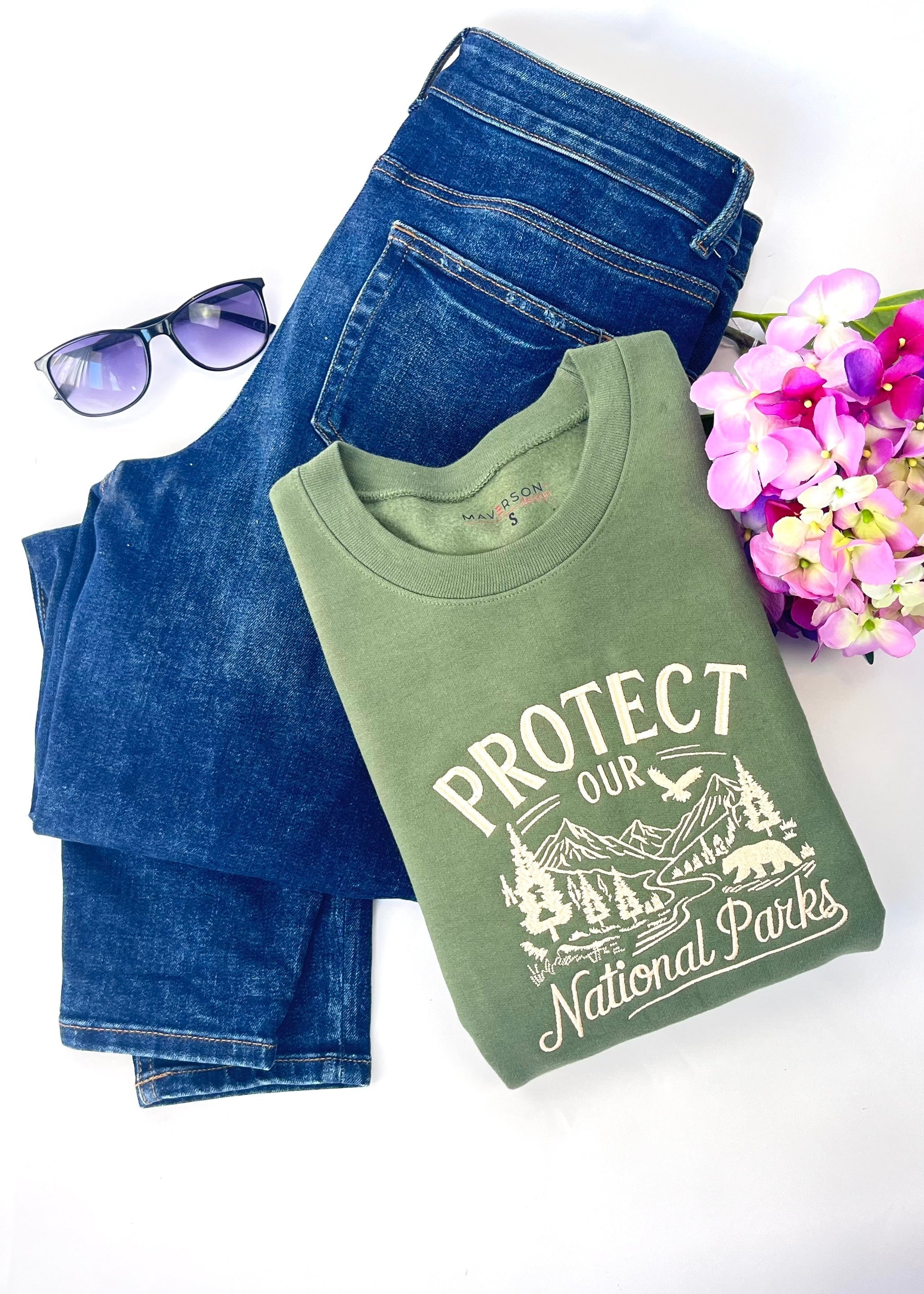 Green t-shirt with 'Protect our National Parks' text, blue jeans, sunglasses, and flowers on a white background