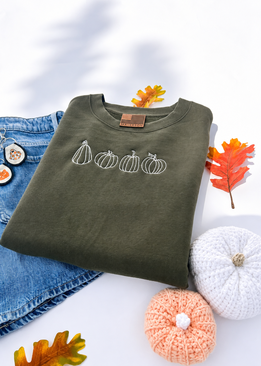 Green sweatshirt with pumpkin design on a white surface with autumn leaves and pumpkins.